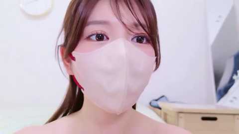 Video of a young Asian woman with light skin, brown eyes, and long brown hair tied back, wearing a white surgical mask. She has a slightly startled expression. The background shows a white wall, a clock, and a wooden chest of drawers.