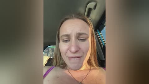 Video of a Caucasian woman with long blonde hair, wearing a purple top, seated in a car. She has a solemn expression, with slightly closed eyes and a slight frown. The background shows the car's interior.