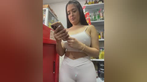 Video of a Latina woman with long dark hair, light skin, wearing a white crop top and high-waisted pants, shopping at a convenience store, checking her phone.