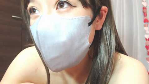 Video of a young Asian woman with long brown hair, wearing a light blue surgical mask, looking upward. She has a fair complexion and is partially visible, with a blurred background featuring a wooden door and a string of red and white beads.