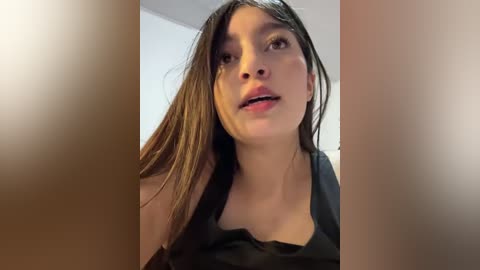 A video of a young woman with long brown hair and light skin, wearing a black top, looking up with a neutral expression, set against a blurred, neutral-toned background.