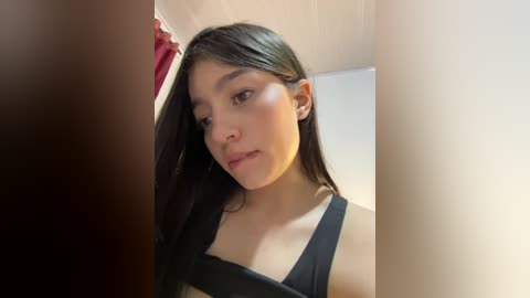 Video of a young Latina woman with long dark hair, wearing a black tank top, standing in a bathroom with beige walls and a red curtain.
