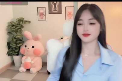 Video of a young East Asian woman with long black hair, light skin, and red lipstick, wearing a light blue button-down shirt. Background features a pink bunny plush, a green potted plant, and framed art on a light-colored wall.