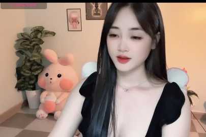 Video of a young East Asian woman with pale skin, long black hair, and red lipstick. She wears a low-cut black dress, standing next to a plush bunny and green plant in a checkered room.