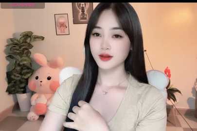 A video of an East Asian woman with long black hair, fair skin, and red lipstick, wearing a beige top, in a cozy room with a pink bunny plush, framed photos, and potted plants.