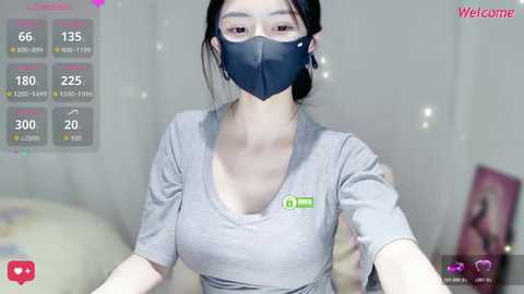 Video of an East Asian woman with fair skin, dark hair in a bun, wearing a blue face mask, gray T-shirt, and a green logo on her chest, in a bedroom setting.