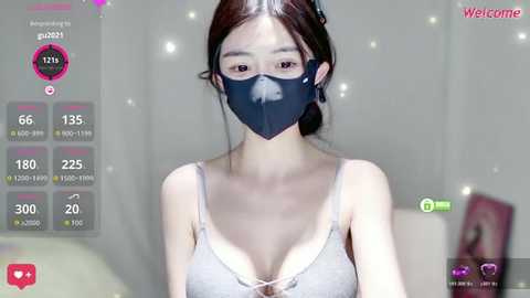 A video of an East Asian woman with light skin and brown hair in a ponytail, wearing a black face mask and a light gray bra. She stands in a dimly lit room with a soft focus background.