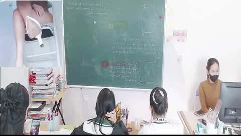 Video of a classroom with two Asian women wearing face masks and black dresses, one on a ladder, one seated at a cluttered desk, and a green chalkboard with equations.