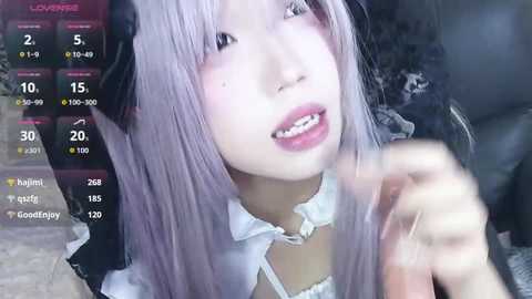 Video of a young, fair-skinned woman with long, light lavender hair and a black bow, wearing a maid outfit, smiling while holding a camera.