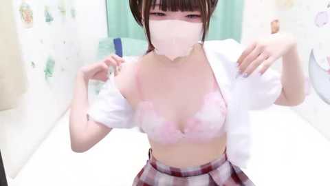 A video of an East Asian woman in a pink lace bra and a white shirt, with a plaid skirt, standing in a bathroom. She has a white mask covering her face, and is adjusting her shirt.