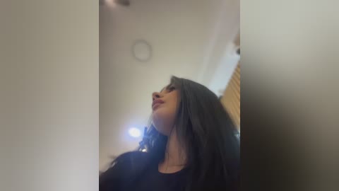 Video of a young woman with long, dark hair, wearing a black top, looking upward with a serene expression. Background features a minimalist, off-white room with a circular clock on the ceiling.