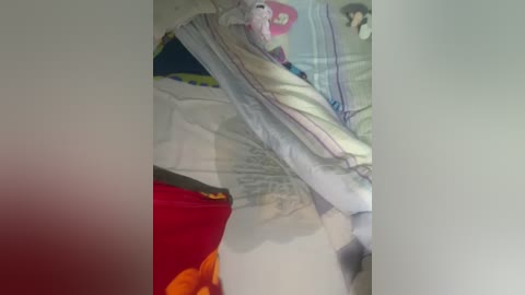 A video captures a messy bedroom with a pink duvet, a red blanket, and a beige pillow. A partially visible person lies on the bed, wearing a white nightgown. The background is blurry, with a white wall.