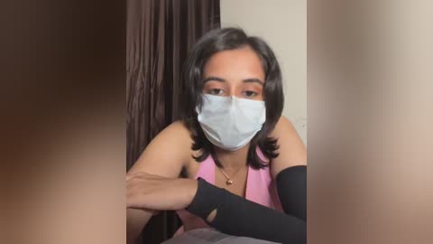 Video of a South Asian woman with medium skin tone, wearing a pink sleeveless top, black elbow-length gloves, and a light blue face mask, seated indoors with a brown curtain in the background.