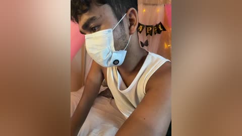 Video of a young South Asian man with short, curly hair, wearing a white tank top and a white face mask, lying on a bed with a pink background.