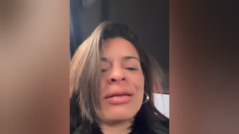 A video of a woman with long, straight, dark hair and light skin, wearing a black jacket, smiling slightly with closed eyes.