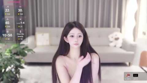 Video of a young East Asian woman with long black hair, fair skin, and a slender physique, topless, in a modern living room with a gray sofa and a potted plant.