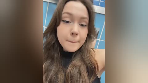 Video of a young woman with long, wavy brown hair, wearing a black sleeveless top. She has light skin, a straight nose, and a slight smile. The background features blue-tiled walls and a blurred, warm-toned foreground.