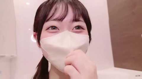 Video of an Asian woman with straight dark hair, wearing a white face mask, looking forward, holding the mask, in a neutral indoor setting with white and brown walls.