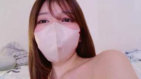Video of an Asian woman with long brown hair, wearing a white face mask, in a bedroom with white sheets and a floral patterned pillow.
