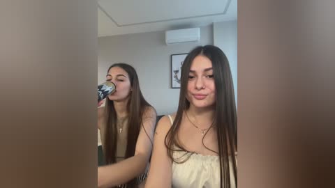 Video of a young woman with long brown hair, light skin, wearing a strapless white top, drinking from a plastic cup, in a modern room with neutral walls, framed artwork, and an air conditioner.