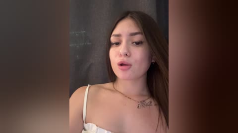 Video of a young East Asian woman with long brown hair, fair skin, and wearing a white lace bra. She has a neutral expression, with a small heart tattoo on her neck. The background is dimly lit, featuring a dark curtain.