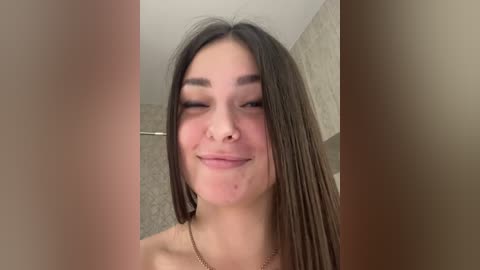 A young woman with long brown hair, light skin, and a nose piercing smiles in a bathroom with beige tiles.