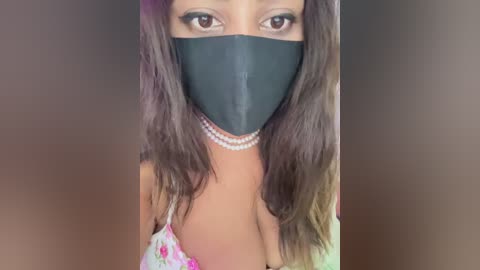 Video of a Black woman with long, wavy hair wearing a black face mask, floral-patterned top, and pearl necklace. The background is a blurred gradient of brown tones.