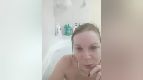 A video of a topless, fair-skinned woman with wet, brown hair, standing in a white bathtub, with a blurred man in the background.