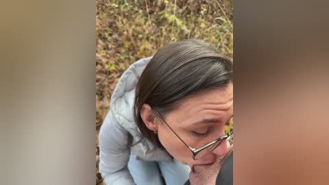 Media: A video of a woman with glasses, light-colored hair, and a light jacket, crouching on a grassy field with autumn foliage in the background. She appears to be focused on something on the ground.