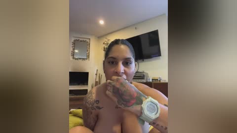 A video of a topless woman with dark hair, tattoos, and an Apple Watch, sitting on a bed, biting her finger, in a cozy room with a TV and mirror.