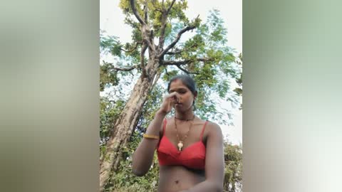 Video of a dark-skinned woman with medium build, wearing a red bra and gold necklace, standing in a forest with a tall, leafy tree in the background.