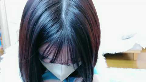 Media: Video of a person with long, straight, dark brown hair partially covering their face, wearing a white surgical mask, standing indoors with a reflective surface and a white background.