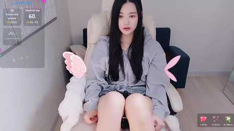 Video of a young East Asian woman with long black hair, sitting in a plush armchair, wearing a grey hoodie and black shorts, holding a pink shoe, in a minimalist room with a TV screen displaying game statistics.