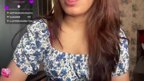 Video of a woman with long, wavy brown hair and a light brown complexion, wearing a blue floral top. Background features a floral-patterned wall and a pink object in the lower left.