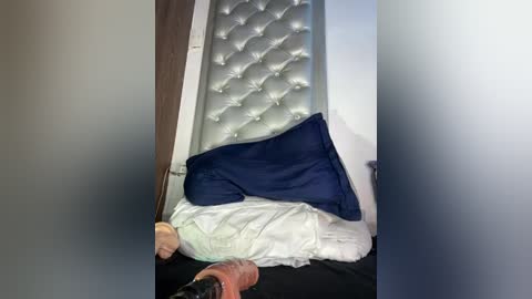 Video of a bed with a tufted, silver headboard, a white pillow, and a navy blue pillow, partially covered by a white sheet, in a dimly lit room with a blurred door frame.