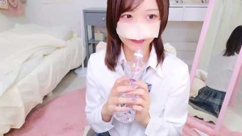 Video of an Asian woman with brown hair, wearing a white shirt, holding a clear plastic cup, in a pink room with a bed and white furniture.