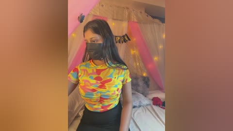 Video of a young woman with long black hair, wearing a colorful, form-fitting top, black face mask, and black skirt, in a cozy bedroom with pink and white canopy, fairy lights, and a bed with white sheets.