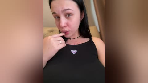 Video of a young woman with pale skin and long black hair, wearing a black tank top with a heart design. She has a neutral expression, biting her finger, and is standing in a dimly lit room with beige walls.