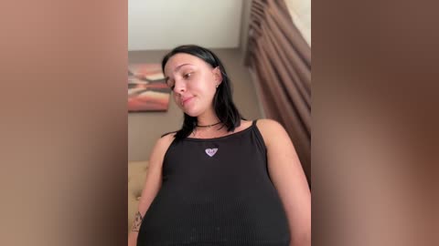 A video of a young woman with fair skin, black hair, and a black tank top with a purple heart, standing in a dimly lit room with beige walls and brown curtains.