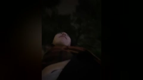 A dimly lit video of a person lying on their back, with their head tilted upward, looking serene. The background is dark and indistinct, creating a mysterious, dream-like atmosphere.