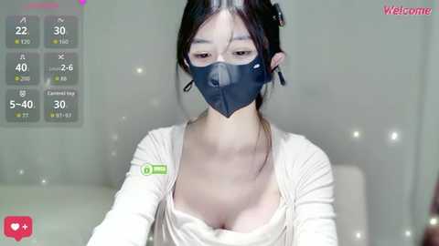 Video of a young Asian woman with fair skin, wearing a black face mask, white long-sleeve top, and a low-cut neckline, sitting in front of a light-colored background.