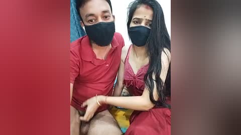 Video of a couple in casual attire, both wearing black face masks, sitting closely together; the woman has long dark hair and wears a red top.