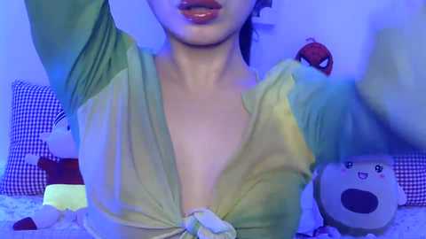 Video of a young woman with light skin, dark hair, and glossy lips, wearing a low-cut, green top with a knot in front. Background features a blue-tinted room with plush toys, including a large bear.