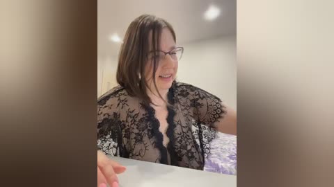 A video of a smiling woman with glasses, light skin, and shoulder-length brown hair, wearing a black lace robe, sitting at a table.