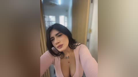 Video of a Latina woman with long black hair, wearing a low-cut pink top, leaning against a door frame in a hallway.