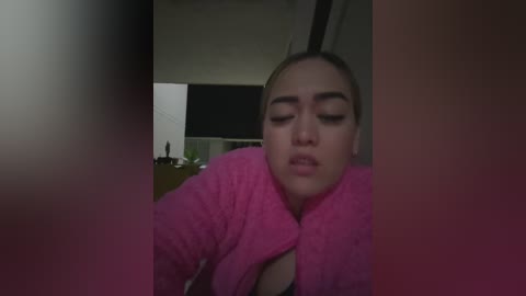 Video of a woman with light skin, dark hair tied back, wearing a pink fluffy bathrobe, looking tired and emotional, blurred background with a lamp and a black wall.