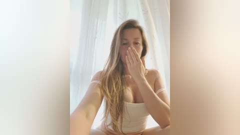 Video of a young woman with long, light brown hair, partially covering her face with her hands. She is wearing a white off-shoulder crop top, sitting in front of sheer white curtains. The image has a soft, ethereal quality.