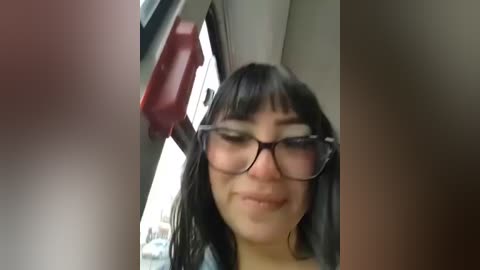 Video of a woman with straight black hair, wearing black-framed glasses and a gray shirt, smiling in a car. The background shows a red seatbelt and a blurred interior.