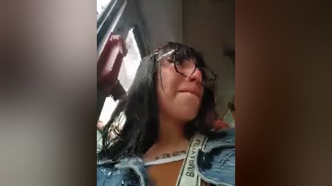 A video of a woman with shoulder-length dark hair, wearing glasses, a blue denim jacket, and a white strap, standing in a dimly lit room with a window in the background.