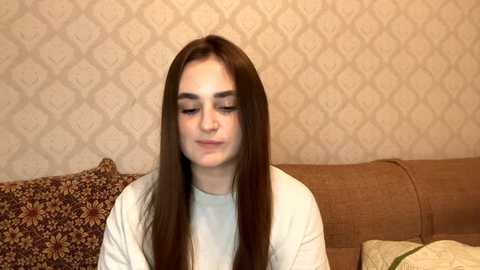 Video of a young woman with long brown hair, wearing a white shirt, seated on a brown couch with floral-patterned cushions against a beige, diamond-patterned wallpaper background.